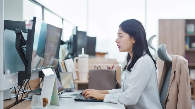 A young professional woman sitting at a workstation with two computer monitors in a bright modern open-plan office environment.
