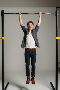 Young man with scared expression hangs from metal bar. Arms raised in panic, holding tightly to bar. Wears jeans, patterned shirt over white t-shirt, with brown boots. Background plain gray studio