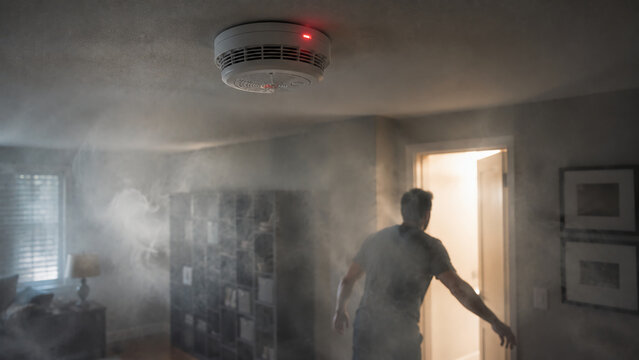 A person enters a smoke-filled room, highlighting a potential fire hazard and the importance of a smoke detector installed on the ceiling.