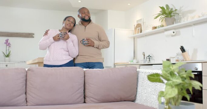 African American couple standing by sofa in open kitchen, holding mugs and watching off-camera