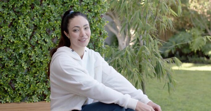 Woman wearing white hoodie sitting on bench in yard responding to camera, straightening and smiling