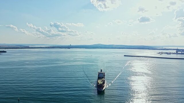 A cargo ship moves through a wide body of water with gentle waves under a clear sky. The sun shines brightly as it approaches the port area in the background.