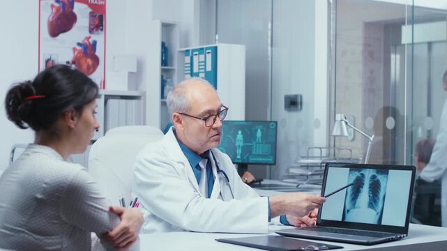 Senior doctor discusses lung X ray results with a young female patient, explaining possible pneumonia, cancer risk and specialist examination, with scan review and specialist medical advice.