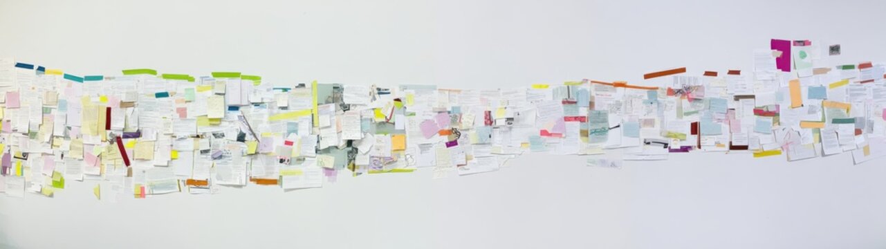 An ultra-wide photograph of a long, chronological paper timeline of forty overlapping, multi-colored sticky notes and other paper scraps, attached to a seamless white background