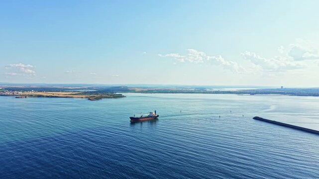 A cargo ship navigates through still waters close to a coastal town. Fields and hills are seen in the distance under a bright sky. It is a sunny afternoon with few clouds.