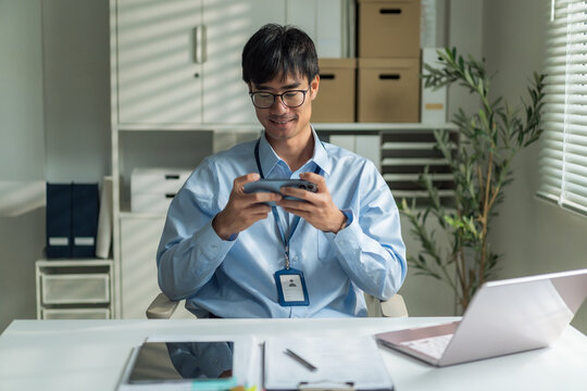 Young office worker wearing glasses smiling happily while relaxing and playing mobile game on smartphone at desk.