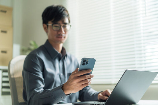 A young businessman wearing glasses is using his smartphone and working on his laptop at a desk in a sunlit office. Choose a focal point.
