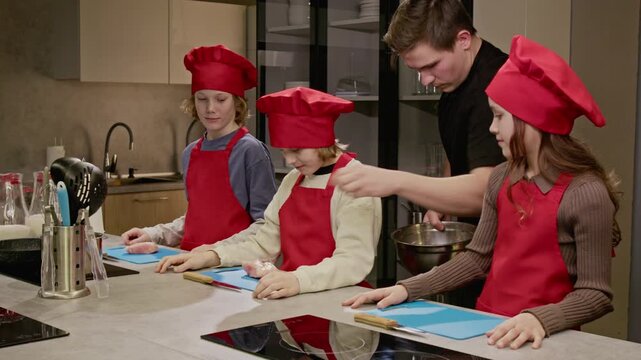 Caucasian male chef distributing raw chicken to Caucasian children during culinary class in modern kitchen. Ideal for promoting children cooking workshops and educational programs