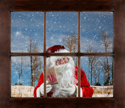 Santa Claus holding a present looking through a six pane wood window with a winter scene in the bacckground