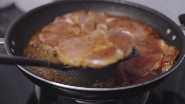 Marinated pork steaks sizzling in a black non-stick skillet on a stovetop. Close up of juicy meat pieces simmering in savory brown sauce with a spatula.