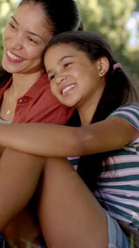 Vertical video: Leaning mom and girl smiling after laugh on sunlit grass in park, pendant, hair tie
