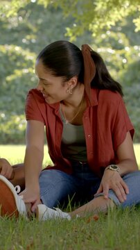 Vertical video: Spotting loose lace, mother tying white shoe on grass in park in rust-red shirt