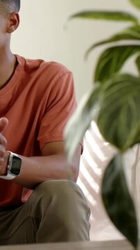 Vertical video: Camera panning man in coral tee gesturing, talking on sofa, showing watch and plant
