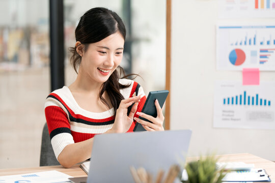 Happy young Asian businesswoman using smartphone while working on laptop in modern office.