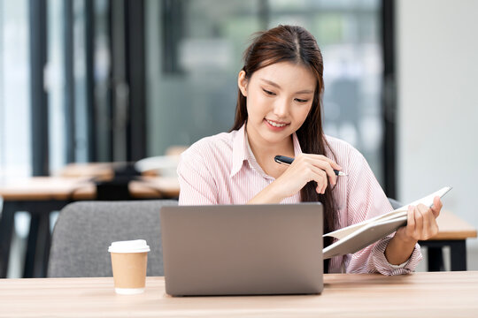 Young Asian businesswoman working on laptop and holding notepad in modern office.