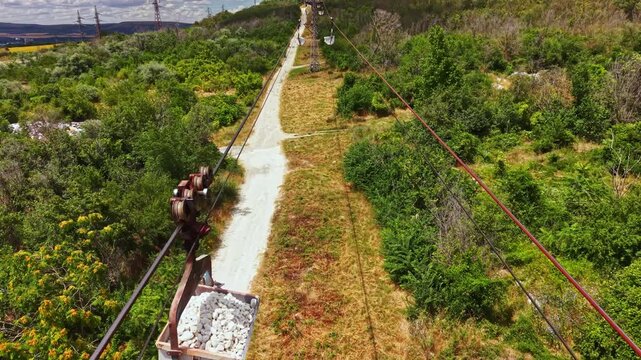 Workers transport rocks using a cable system in a forested area. The path shows cleared land and greenery around. This scene takes place on a sunny day in summer.