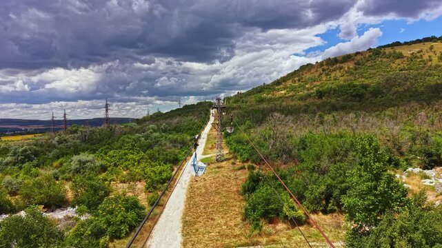 A group of zip liners glide across a course on a hillside. The course runs along a path with trees on both sides. The sky shows clouds indicating possible weather changes.