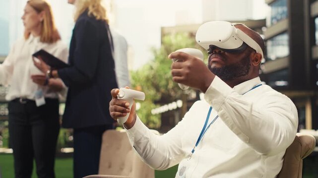 Black business employee in VR glasses uses handheld controllers sitting outdoor with coworkers at whiteboard in blurred background, doing interactive simulation, for business planning.
