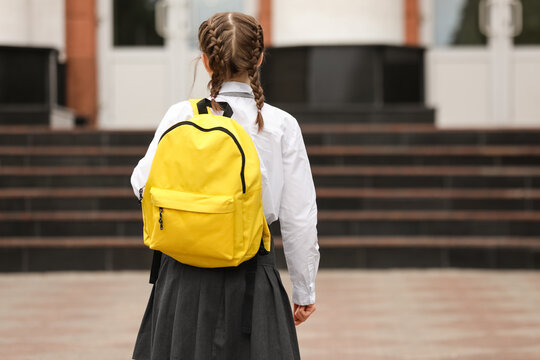 Young schoolgirl with braided hair and a bright yellow backpack walking towards a school building entrance, the student wears a white shirt and gray skirt, a typical school uniform