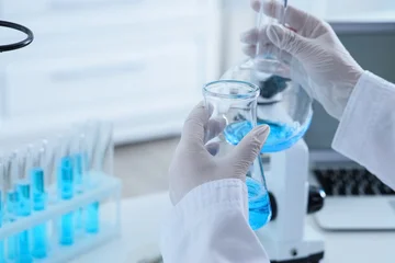 Scientist in white lab coat and gloves holding two beakers with blue liquid in a laboratory setting, the scene takes place in a modern laboratory, with additional test tubes in a rack © Pixel-Shot
