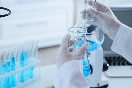 Scientist in white lab coat and gloves holding two beakers with blue liquid in a laboratory setting, the scene takes place in a modern laboratory, with additional test tubes in a rack