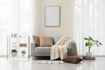 Cozy living room interior with a gray sofa, throw pillows, blanket, and a blank picture frame on the wall © Pixel-Shot