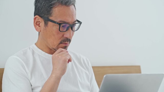 ベッドでノートPCを使うシニア男性。パソコン、仕事、DX、リラックス、ワーケーション、テレワーク