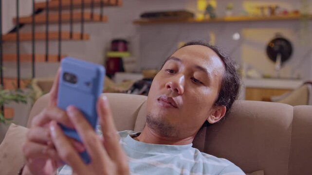 A man leans back on a sofa while using his smartphone, capturing a relaxed home lifestyle, mobile browsing, and everyday digital technology use.