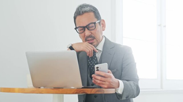 スマホとノートPCを使いながら悩むミドル男性ビジネスパーソン。不満、疑問、アプリ、失敗、残念、SNS