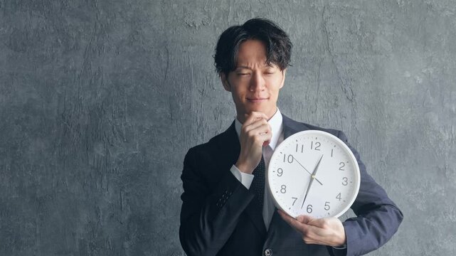 時計を持って悩む若い男性ビジネスパーソン。時間、〆切、スケジュール、不安、心配、忙しい、多忙、疑問