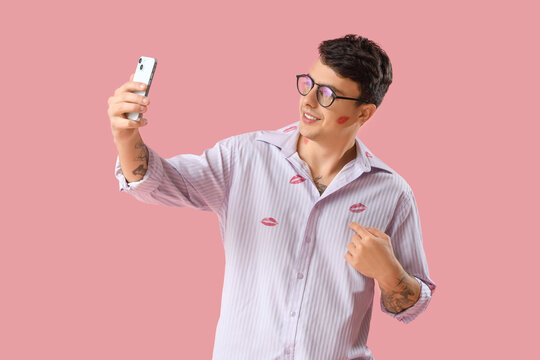 Young man with kiss marks on his shirt and mobile phone against pink background