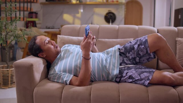 A man lies on a sofa while browsing social media on his smartphone, capturing a relaxed home lifestyle, mobile screen time, and everyday digital entertainment.