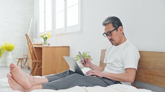 ベッドでノートPCを使うシニア男性。パソコン、仕事、DX、スマホ、アプリ、ワーケーション、テレワーク