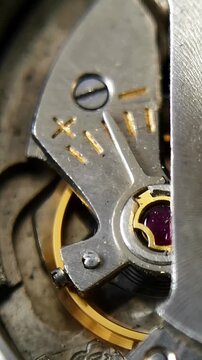 Vertical extreme close up of a vintage watch movement showcasing the balance cock plate with engraved plus and minus adjustment symbols, ruby jewel, and a pulsing golden balance wheel.