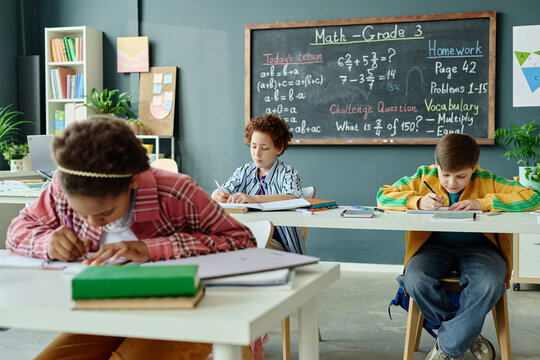Wide shot of diverse school kids writing math exercises at desks during classroom lesson, showing focused learning and academic progress. Ideal for education, tutoring, curriculum marketing