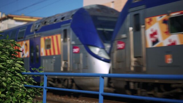 Moving train passing behind a blue railing, with colorful exterior graphics and greenery in the foreground, creating a dynamic urban travel scene