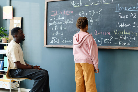 African American schoolgirl solving math problem on classroom chalkboard while male teacher observing, showing guided learning and academic support. For education campaigns and tutoring ads