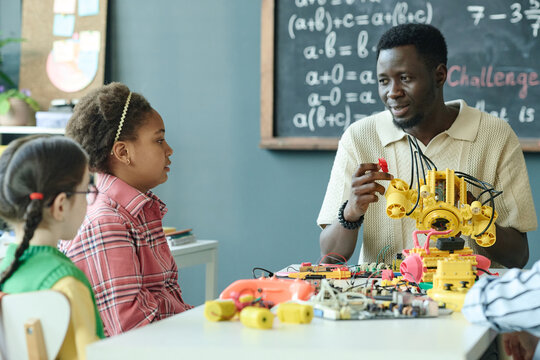 Medium shot of Black male teacher guiding diverse children in robotics lesson, demonstrating engineering model in classroom. Ideal for STEM education, coding camp, school marketing