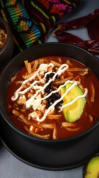 Mexican tortilla soup in a black ceramic bowl with avocado, cheese and pasilla chili