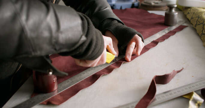 Footwear manufacturing. Shoes factory. Unrecognizable craftsman hands using stationery knife metal ruler cutting material at workspace.