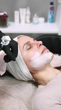 Vertical video. Peeling mask. Skin treatment. Professional beautician using brush applying moisturizing cream on female face in beauty salon.