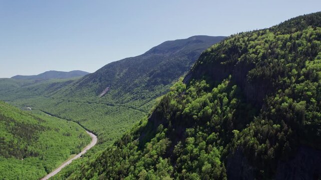 Amazing drone fly of lush green mountains environment near winding road running through the deep White Mountain National Forest valley under clear blue sky, New Hampshire, United States