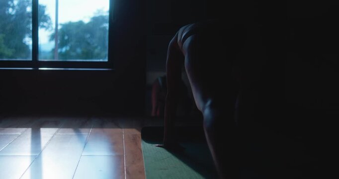 Dark indoor silhouette of a woman performing slow bodyweight floor exercise near a window on a yoga mat at home.