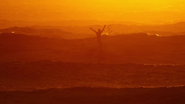 Stoked kiteboarder throws hands in air in vivid golden sunset light, telephoto