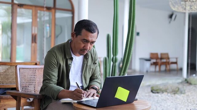 Asian businessman doing business research using laptop writing in notebook in cafe. Business and modern lifestyle concept.