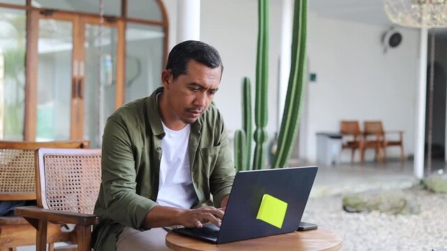 Overworked concentrated Asian copy-writer wearing shirt working on new articles using laptop computer, leaning his elbow on cafe table