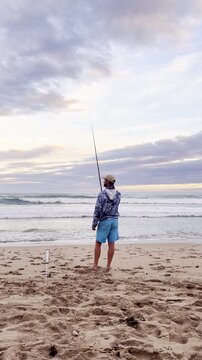 Barefoot avid fisherman fishing from sandy beach at sunset. Rear vertical