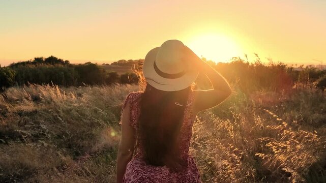 A woman in a hat gracefully walks through a field of golden wheat, bathed in the warm light of a beautiful sunset. Her long hair flows gently in the breeze, capturing the serene beauty of nature and g