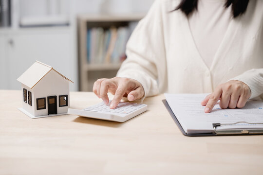 Investment analysis for home property mortgage loan shows female hands using calculator and reviewing financial contract paper next to small house model on office desk