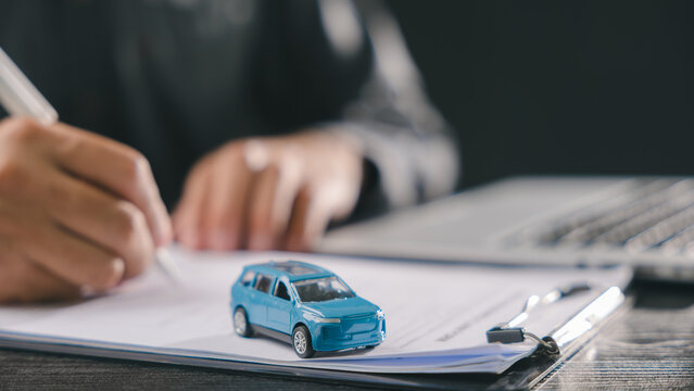Buyer signs car loan purchase finance vehicle contract document, securing insurance and ownership with small blue toy model on clipboard while working near laptop in office